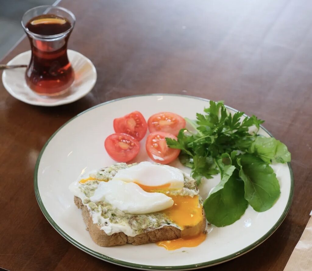 A healthy breakfast plate at Ceviz Ev Yapımı featuring avocado toast topped with runny poached eggs, cherry tomatoes, greens, and Turkish tea.