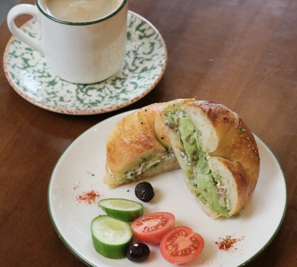 A breakfast setting at Ceviz Ev Yapımı with a filled bagel sandwich (avocado/cream cheese), cucumbers, olives, and a cup of frothy coffee.