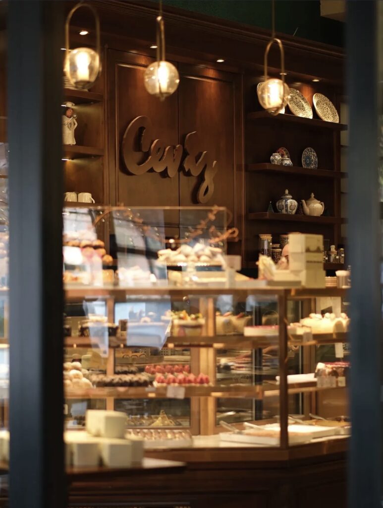 View through the window into Ceviz Ev Yapımı, showing the wooden logo wall, pendant lights, and a refrigerator full of fresh cakes.