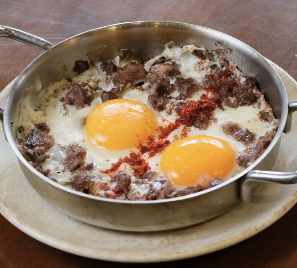 Close-up of a copper pan at Ceviz Ev Yapımı containing fried eggs cooked with savory meat pieces and sprinkled with red paprika.