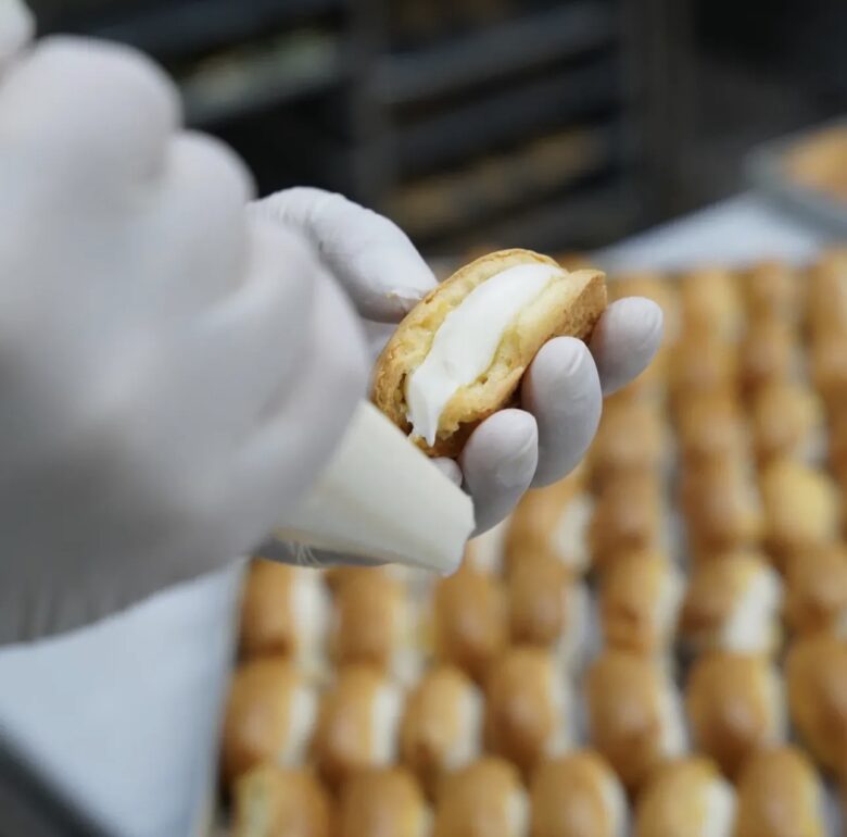 Close-up shot of hands in gloves piping fresh white cream into a pastry shell at the Ceviz Ev Yapımı kitchen.