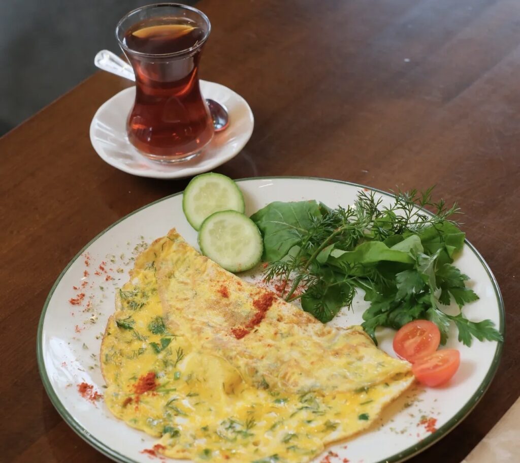 A light breakfast plate at Ceviz Ev Yapımı featuring a yellow omelet mixed with fresh green herbs, served with a side of arugula and tomatoes.